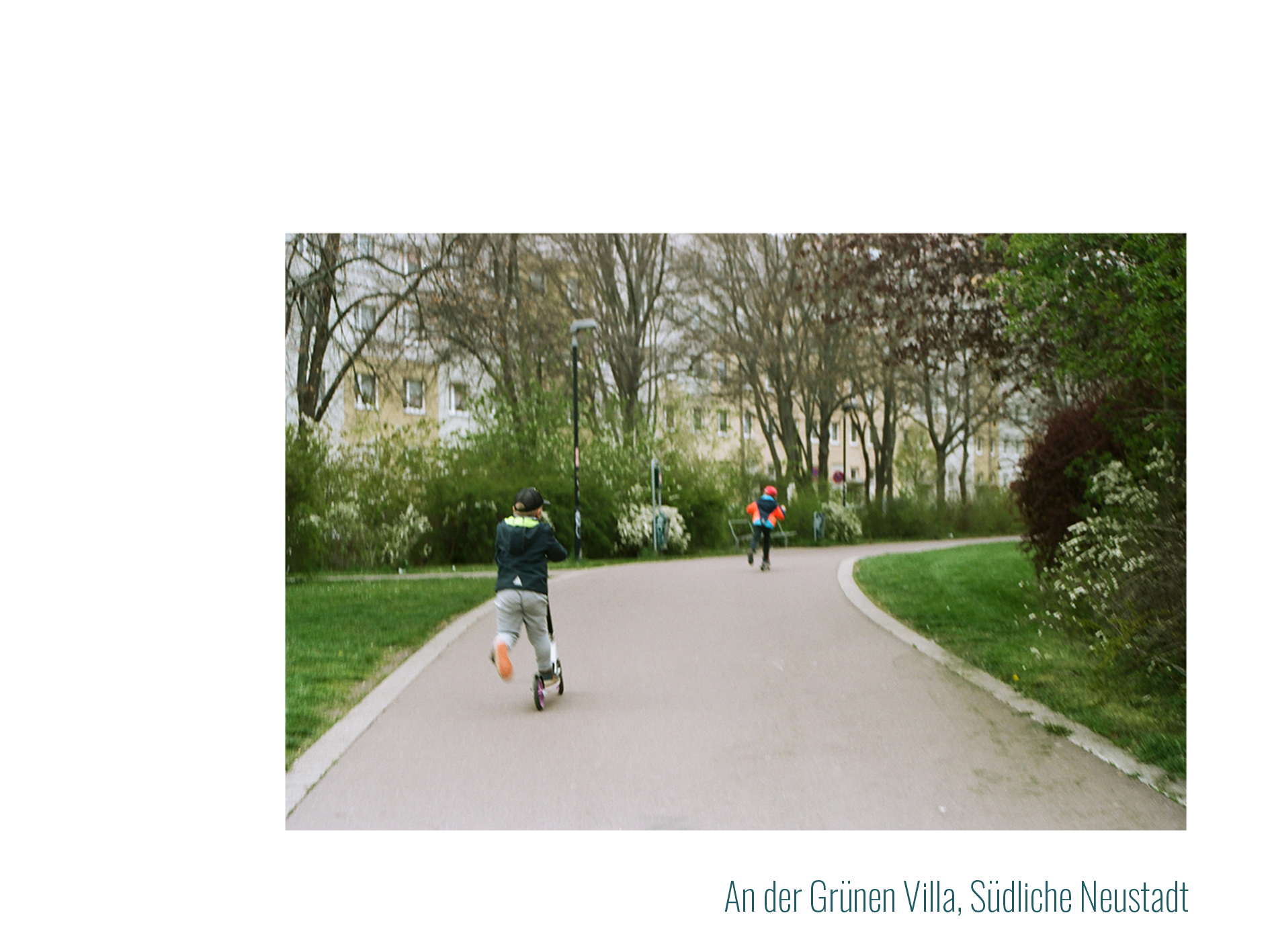 Kulturzentrum Grüne Erde:
            Zwei kleine Jungs fahren auf Rollern einen Gehweg im Park entlang.
            Sie sind von hinten und in Bewegung fotografiert.