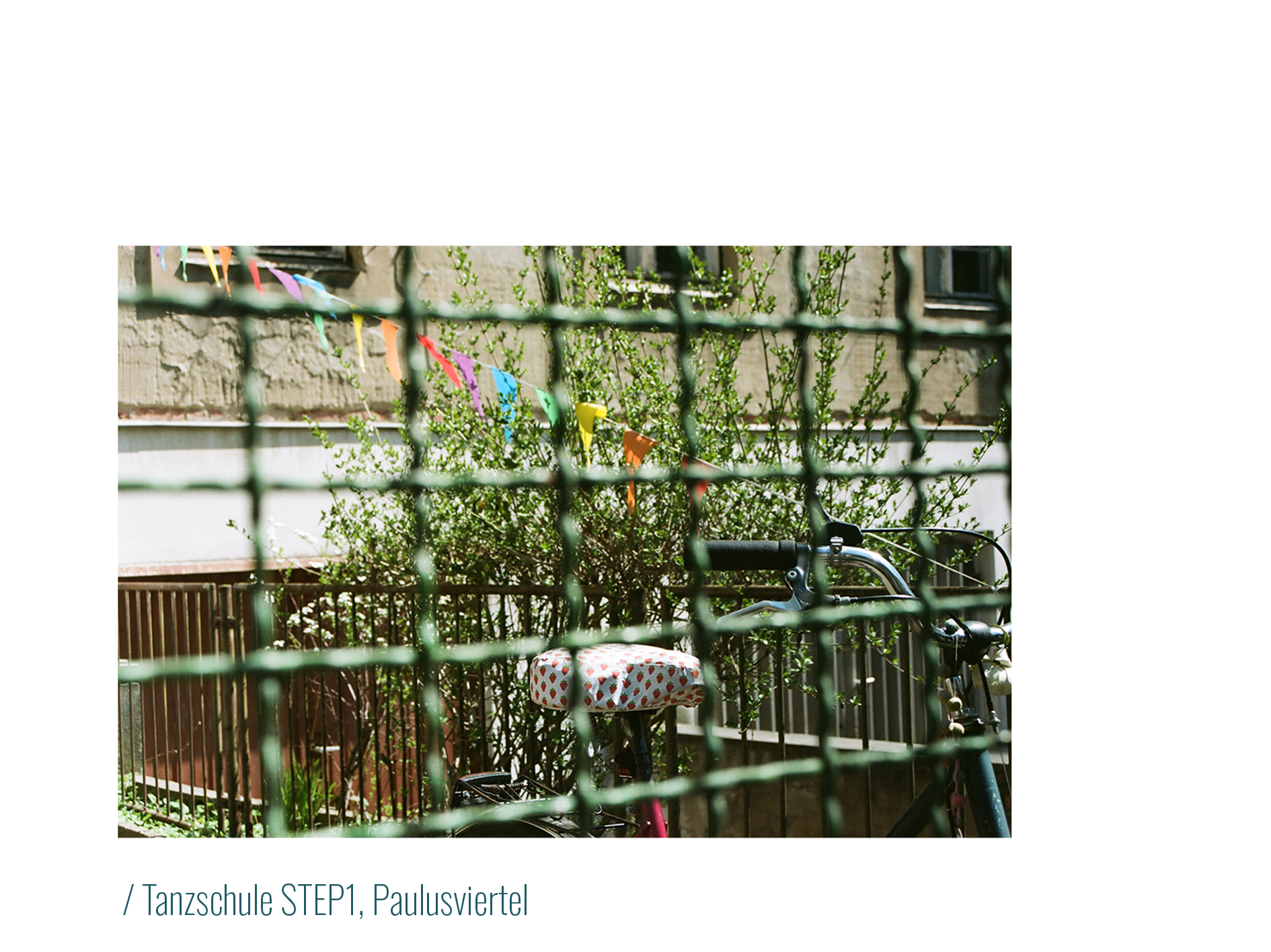 In Kontrast zu Tanzschule STEP1, Paulusviertel:
            Durch einen Zaun blickt man auf ein Fahrrad, auf dessen Sattelschutz kleine rote Erdbeeren abgebildet sind.
            Im Hintergrund weht eine bunte Girlande mit Fähnchen. 